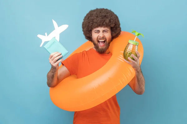 Man with Afro hairstyle standing with orange rubber ring, holding passport document and airplane mockup, drinking cocktail, rejoicing travel tour. Indoor studio shot isolated on blue background.