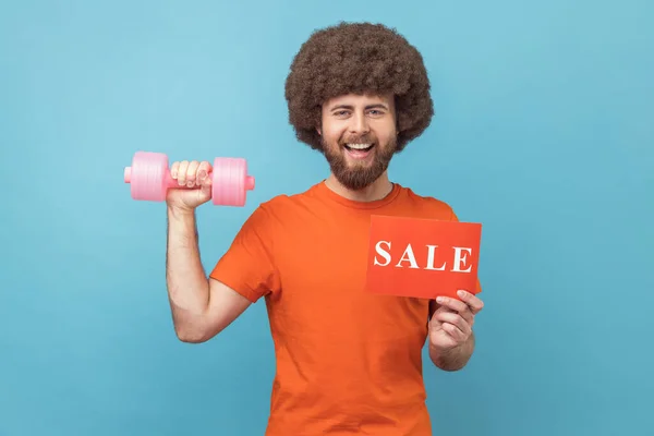 Portrait of satisfied man with Afro hairstyle wearing orange T-shirt holding dumbbell and card with sale inscription, sales for fitness workout. Indoor studio shot isolated on blue background.