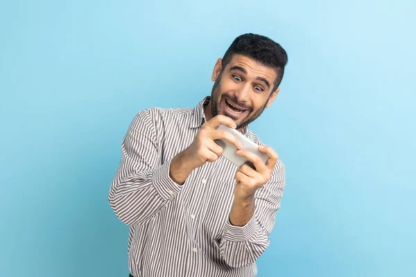 快乐的大胡子商人用快乐积极的表情使用手机 在手机上玩电子游戏 穿着条纹衬衫 室内拍摄被蓝色背景隔离 — 图库照片