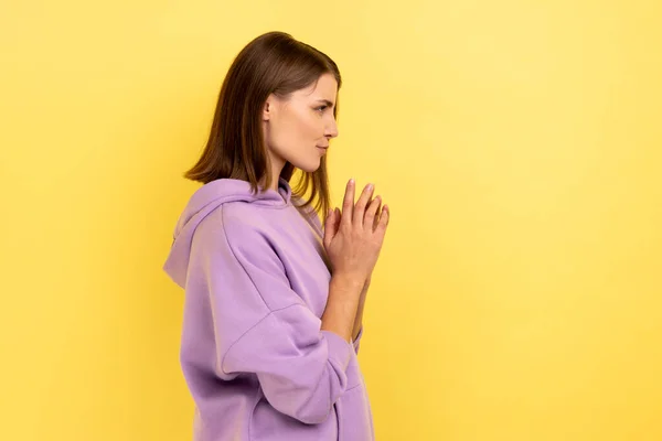 敬虔な狡猾な若い女性の側のビューを手を握りしめ 不思議なことに笑みを浮かべて 悪のいたずら 紫色のパーカーを着て計画 黄色の背景に隔離された屋内スタジオショット — ストック写真
