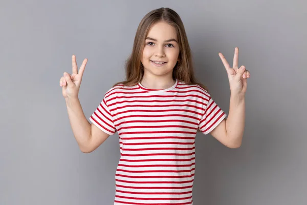 カメラ 成功または達成で勝利サインをジェスチャーストライプTシャツを身に着けている肯定的な楽観的な愛らしい少女の肖像画 灰色の背景に隔離された屋内スタジオショット — ストック写真