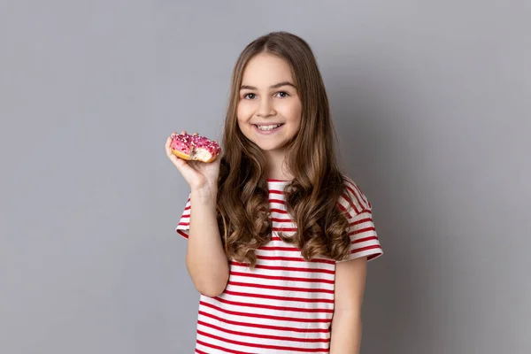 縞模様のTシャツを着た幸せな愛らしい少女の肖像画おいしい甘いドーナツとカメラに本当に笑顔 灰色の背景に隔離された屋内スタジオショット — ストック写真