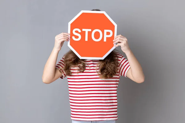 Little girl in striped T-shirt covering face with Stop symbol, anonymous woman holding red traffic sign stop, warning of danger, restriction and limits. Indoor studio shot isolated on gray background.