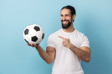 Beyaz tişörtlü, sakallı, elinde futbol topunu gösteren ve dişlek tebessümle kameraya bakan mutlu bir adamın portresi. Mavi arka planda kapalı stüdyo çekimi.