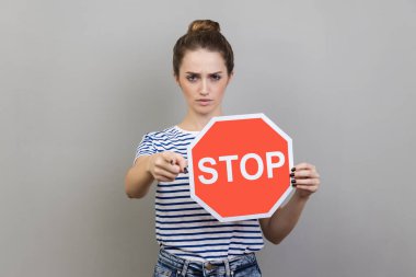 Bun saç stili, çizgili tişört giyen, elinde kırmızı dur işareti taşıyan ciddi patronluk taslayan kadının portresi katı bir ifadeye sahiptir. Gri arka planda izole edilmiş kapalı stüdyo görüntüleri.