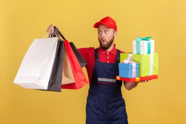 Şaşırmış sakallı kuryenin portresi. Elinde bir sürü hediye kutusu ve alışveriş çantasıyla kapı kapı dolaşıyordu. Sarı arka planda kapalı stüdyo çekimleri.