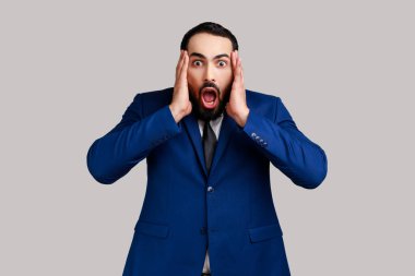 Şaşırmış sakallı adam kameraya ağzı açık bakıyor, avuç içlerini kaldırıyor, şok edici haberlerden etkileniyor, resmi tarz takım elbise giyiyor. Gri arka planda izole edilmiş kapalı stüdyo görüntüleri.