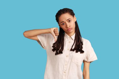 Siyah rastalı hoşnutsuz kadın onaylamadığını gösteriyor, baş parmağını aşağıda tutuyor, hoşnutsuzluğunu ifade ediyor, hoşnutsuzluğunu ifade ediyor, beyaz gömlek giyiyor. Mavi arka planda kapalı stüdyo çekimi.