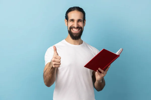 一个积极快乐的男人 留着胡子 身穿白色T恤 竖起大拇指 举手投足看书 喜欢类型和情节 室内拍摄被蓝色背景隔离 — 图库照片