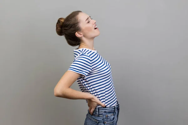 Back, kidney or spine pain. Portrait of woman wearing striped T-shirt standing and endure pain on back, frowning face from pain. Indoor studio shot isolated on gray background.