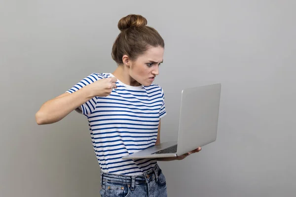 Portrait of aggressive unhappy woman in striped T-shirt having bad mood are ready to punch a worker through a webcam with raised fist with angry face. Indoor studio shot isolated on gray background.