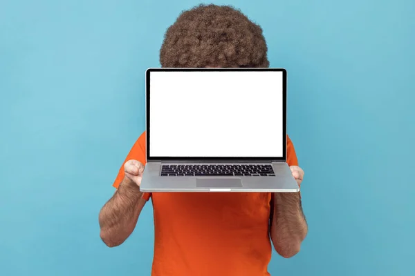 白い空のディスプレイでノートパソコンで顔を隠すオレンジのTシャツを着てアフロの髪型を持つ無名の男 プロモーションテキストのためのコピースペース 青い背景に隔離された屋内スタジオショット — ストック写真