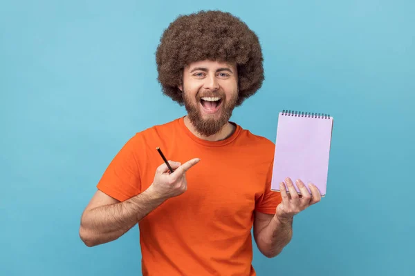 Write reminder to-do list into notebook. Man with Afro hairstyle showing empty paper page, template mock up for creative idea, advertising text. Indoor studio shot isolated on blue background.