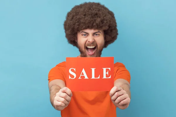 Portrait of excited crazy man with Afro hairstyle wearing orange T-shirt standing showing sale card, presenting low prices, shopping. Indoor studio shot isolated on blue background.