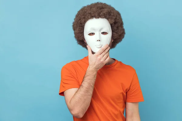 Portrait of man with Afro hairstyle wearing orange T-shirt covering her face with white mask, hiding her real personality, anonymity. Indoor studio shot isolated on blue background.
