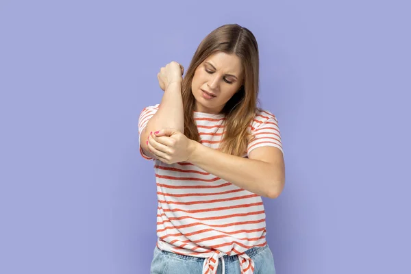 Unhealthy sick blond woman wearing striped T-shirt having trauma after car accident, feels pain in elbow, trapped nerves, rheumatoid arthritis. Indoor studio shot isolated on purple background.