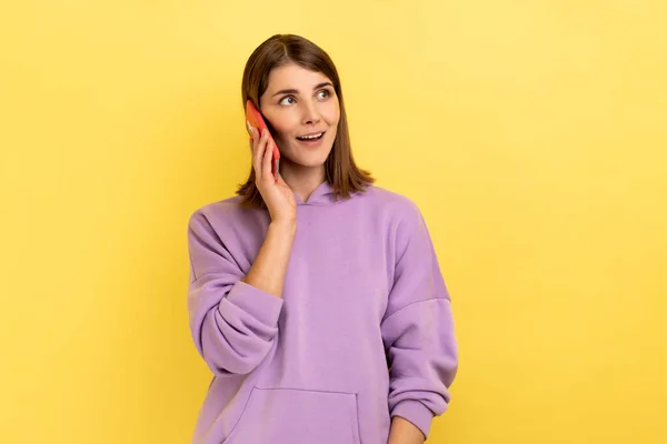 携帯電話で友人と話す陽気な女性の肖像画 離れて見て 楽しく笑顔 楽しい会話をし 紫色のパーカーを着ている 黄色の背景に隔離された屋内スタジオショット — ストック写真