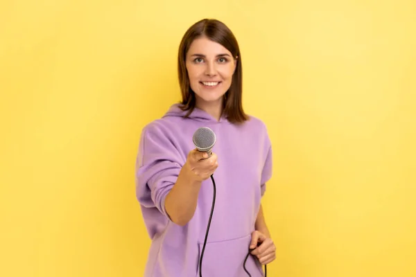 Portrait of satisfied delighted woman with dark hair standing offering microphone, journalist taking interview, wearing purple hoodie. Indoor studio shot isolated on yellow background.