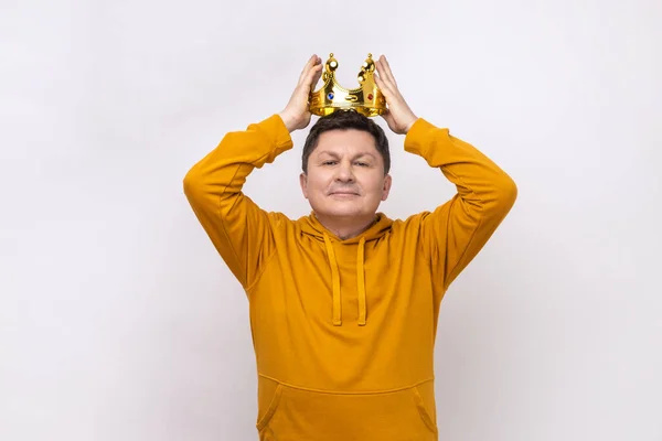 I'm ceo. Man wearing golden crown, imagining promotion at work, looking with arrogance, privileged status, wearing urban style hoodie. Indoor studio shot isolated on white background.