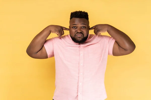 I dont want to listen it. Annoyed man hears loud noise, avoids unpleasant sound, doesnt hear somebody\'s advice, closes ears with index fingers. Indoor studio shot isolated on yellow background.