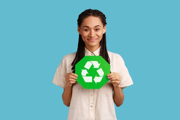 Responsible self confident woman with black dreadlocks holding green recycling sign in hand and seriously looking at camera, thinking green. Indoor studio shot isolated on blue background.