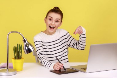 İş yerinde grafik tablet ve dizüstü bilgisayarla oturup kameraya heyecanla bakan zevkli yaratıcı kadın tasarımcı. Sarı arka planda kapalı stüdyo çekimleri.