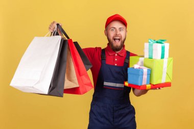 Bir sürü hediye kutusu ve alışveriş çantasıyla kapı kapı dolaşıp kameraya bakan heyecanlı sakallı kuryenin portresi. Sarı arka planda kapalı stüdyo çekimleri.