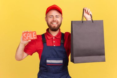 Kameraya bakan mutlu kurye portresi dişlek gülümsemeli, elinde siyah alışveriş çantası ve satılık sözcüğü tutan mavi tulumlu. Sarı arka planda kapalı stüdyo çekimleri.
