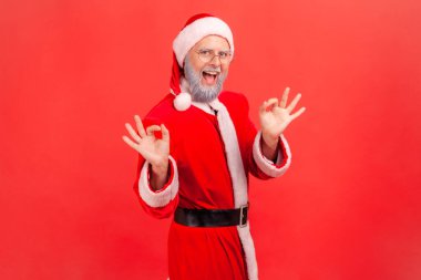 Mutlu yaşlı adam portresi gri sakallı, Noel Baba kostümü giyen, gülümseyen, iyi bir jest yapan, iyi bir işaret gösteren. Kırmızı arkaplanda kapalı stüdyo çekimleri.