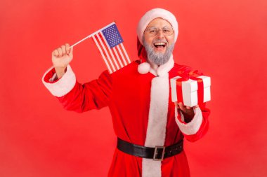 Gri sakallı, Noel Baba kostümü giyen heyecanlı yaşlı bir adamın portresi. Amerikan bayrağını elinde tutuyor ve kış tatillerinde hediye kutusunu veriyor. Kırmızı arkaplanda kapalı stüdyo çekimleri.
