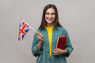 Çekici, zeki, neşeli bir kadının portresi. El kitabı ve İngiliz bayrağı tutuyor. Yurtdışında eğitim kursları veriyor. Gri arka planda izole edilmiş kapalı stüdyo görüntüleri.
