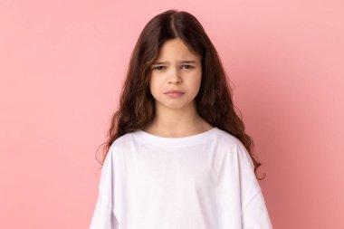 Beyaz tişört giyen üzgün küçük bir kızın portresi negatif duyguları ve üzüntüyü ifade ediyor, kötü bir ruh hali var, üzgün ve mutsuz. Pembe arka planda kapalı stüdyo çekimleri.