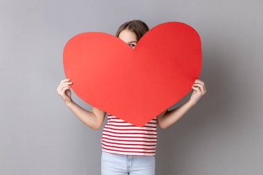 Çizgili tişört giyen küçük sevimli bir kızın portresi büyük kırmızı bir kalbe bakıyor ve olumlu duygular ifade ediyor, aşık oluyor. Gri arka planda izole edilmiş kapalı stüdyo görüntüleri.