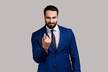 Pozitif gülümseyen yakışıklı sakallı iş adamının portresi. Parmak hareketleriyle arıyor, resmi tarz takım elbise giyip içeri davet ediyor. Gri arka planda izole edilmiş kapalı stüdyo görüntüleri.