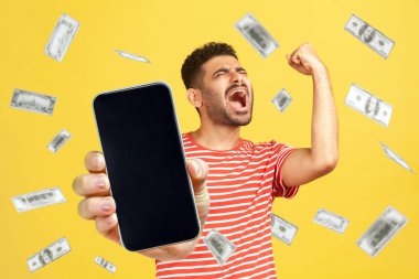 Para yağmuru, kazanan ve zengin. Kırmızı tişört giyen mutlu sakallı adamın portresi elinde akıllı bir telefonla boş ekranı gösteriyor. Sarı arka planda kapalı stüdyo çekimleri.