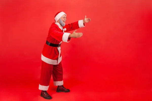 Full length profile shot of elderly man with gray beard wearing santa claus costume reaching hands out, stretching arms to hug someone. Indoor studio shot isolated on red background.