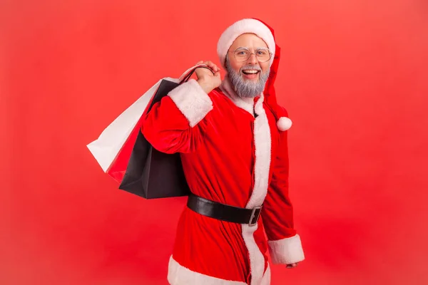 グレーの髭を生やした笑顔の高齢者の肖像は 冬休みのプレゼントを購入し 買い物袋に立ってサンタクラスの衣装を着ています 屋内撮影所は赤の背景に隔離された — ストック写真