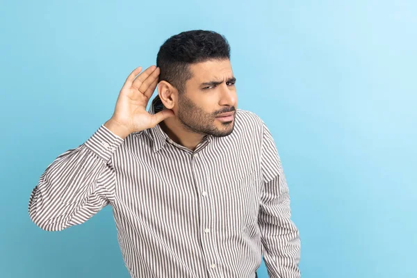 Concentrated nosy businessman with beard holding hand near ear, listening to interesting talks and private secrets, spying, wearing striped shirt. Indoor studio shot isolated on blue background.
