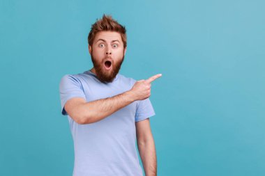 Şaşırmış sakallı adamın portresi fikir sunumu için boş fotokopi alanı gösteriyor, ticari metin, ağzını açık tutuyor. Mavi arka planda kapalı stüdyo çekimi.