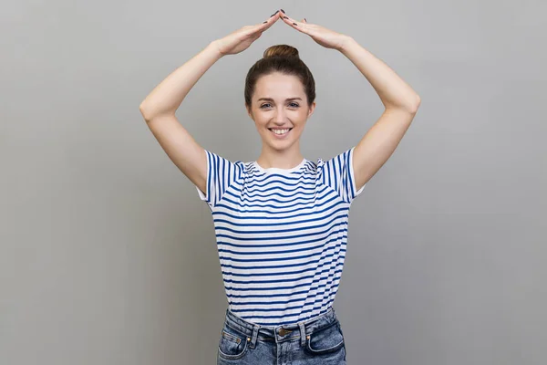 Happy to feel safe. Portrait of woman wearing striped T-shirt showing house roof symbol over head, smiling joyfully, gesture of protection, insurance. Indoor studio shot isolated on gray background.