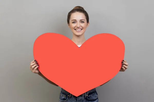 Portrait of happy woman smiling at camera and holding big red heart, symbol of love and affection, expressing i love you and romantic feelings. Indoor studio shot isolated on gray background.