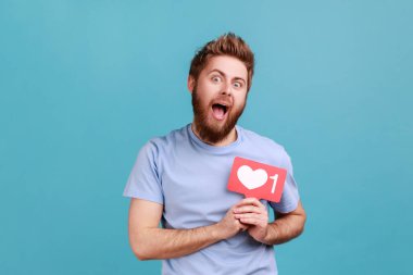 Göğsünün önündeki düğme, emoji sayacı, takipçi tebligatı gibi sosyal medya kalbini tutan heyecanlı sakallı adamın portresi. Mavi arka planda kapalı stüdyo çekimi.
