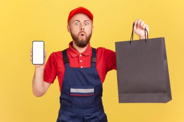 Afallamış teslimatçı portresi mavi üniformalı siyah bir alışveriş çantası ve reklam için boş bir cep telefonu tutuyor. Sarı arka planda kapalı stüdyo çekimleri.