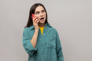 Dedikoducu kız. Ayakta duran, akıllı telefonuyla konuşan, merak eden ve arkadaşlarıyla konuşan sıradan bir ceket giyen bir kadın. Gri arka planda izole edilmiş kapalı stüdyo görüntüleri.