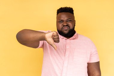 Üzgün, üzgün, sakallı, pembe tişörtlü bir adamın portresi baş parmağını aşağı gösteriyor, olumsuz duygularını ifade ediyor, hoşnutsuzluğunu ifade ediyor, suratını asıyor. Sarı arka planda kapalı stüdyo çekimleri.