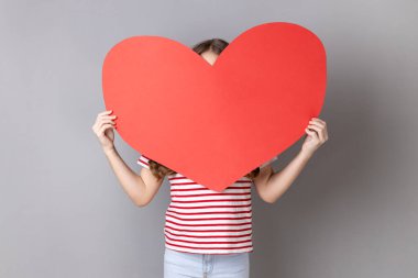 Kimliği belirsiz, çizgili tişört giyen, yüzünü büyük kırmızı kalbin arkasına saklayan küçük kızın portresi duygularını itiraf etmekte tereddüt ediyor. Gri arka planda izole edilmiş kapalı stüdyo görüntüleri.