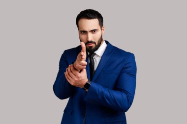 Silah taklit eden ciddi sakallı bir adamın portresi. Parmak tabancalarıyla kameraya ateş ediyor, tehditkar bir şekilde bakıyor ve resmi stil kıyafeti giyiyor. Gri arka planda izole edilmiş kapalı stüdyo görüntüleri.