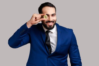 Sakallı iş adamının portresi, altın bitcoin ile gözü kaplayan, dişlek gülümsemeli kameraya bakan, resmi stil kıyafeti giyen. Gri arka planda izole edilmiş kapalı stüdyo görüntüleri.