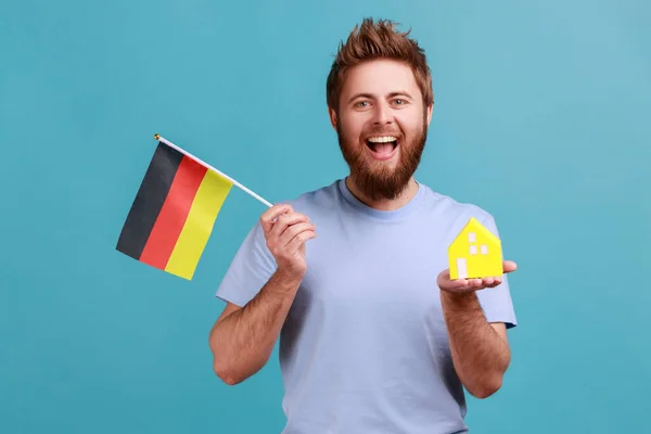 一个积极乐观的英俊大胡子男子手持玩具纸屋和德国国旗 搬到另一个国家的画像 室内拍摄被蓝色背景隔离 — 图库照片