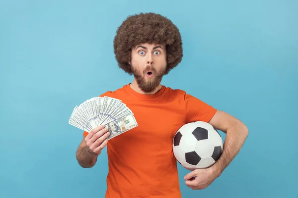 Man with Afro hairstyle wearing orange T-shirt holding soccer ball and hundred dollar bills, looking camera with open mouth, betting and winning. Indoor studio shot isolated on blue background.